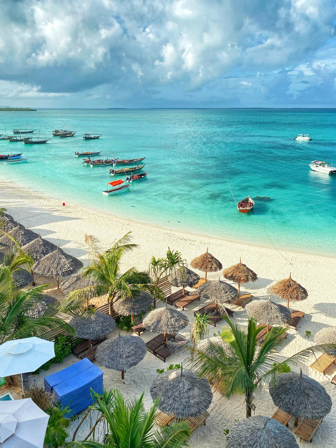 Vista aérea de um resort de praia com guarda-sóis de colmo, espreguiçadeiras e dhows ancorados ao largo