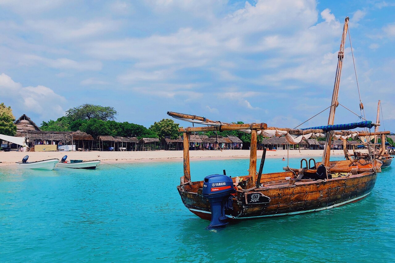 Hölzerne Dhau-Boote liegen in türkisfarbenem Wasser vor einem Strand mit Reetdachhütten und sichtbarem Außenbordmotor