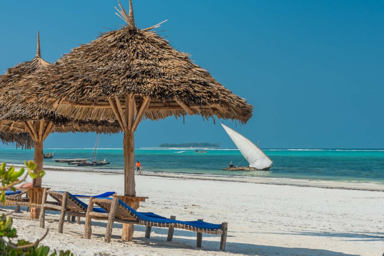 Liegestühle am Strand unter strohgedeckten Sonnenschirmen, im Hintergrund eine segelnde Dhau