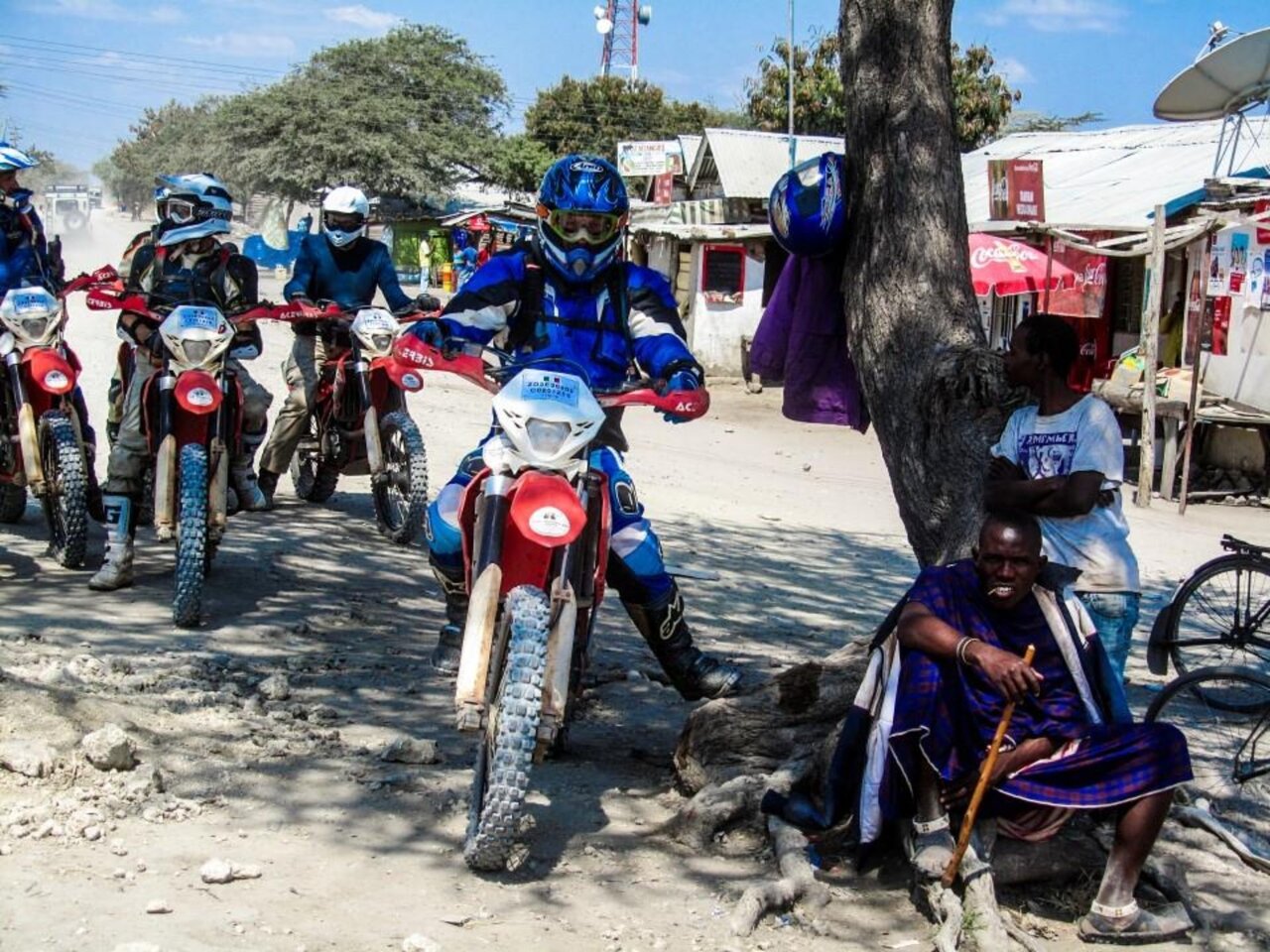 Motoristas detenidos en la calle de un pueblo junto a un hombre masái sentado
