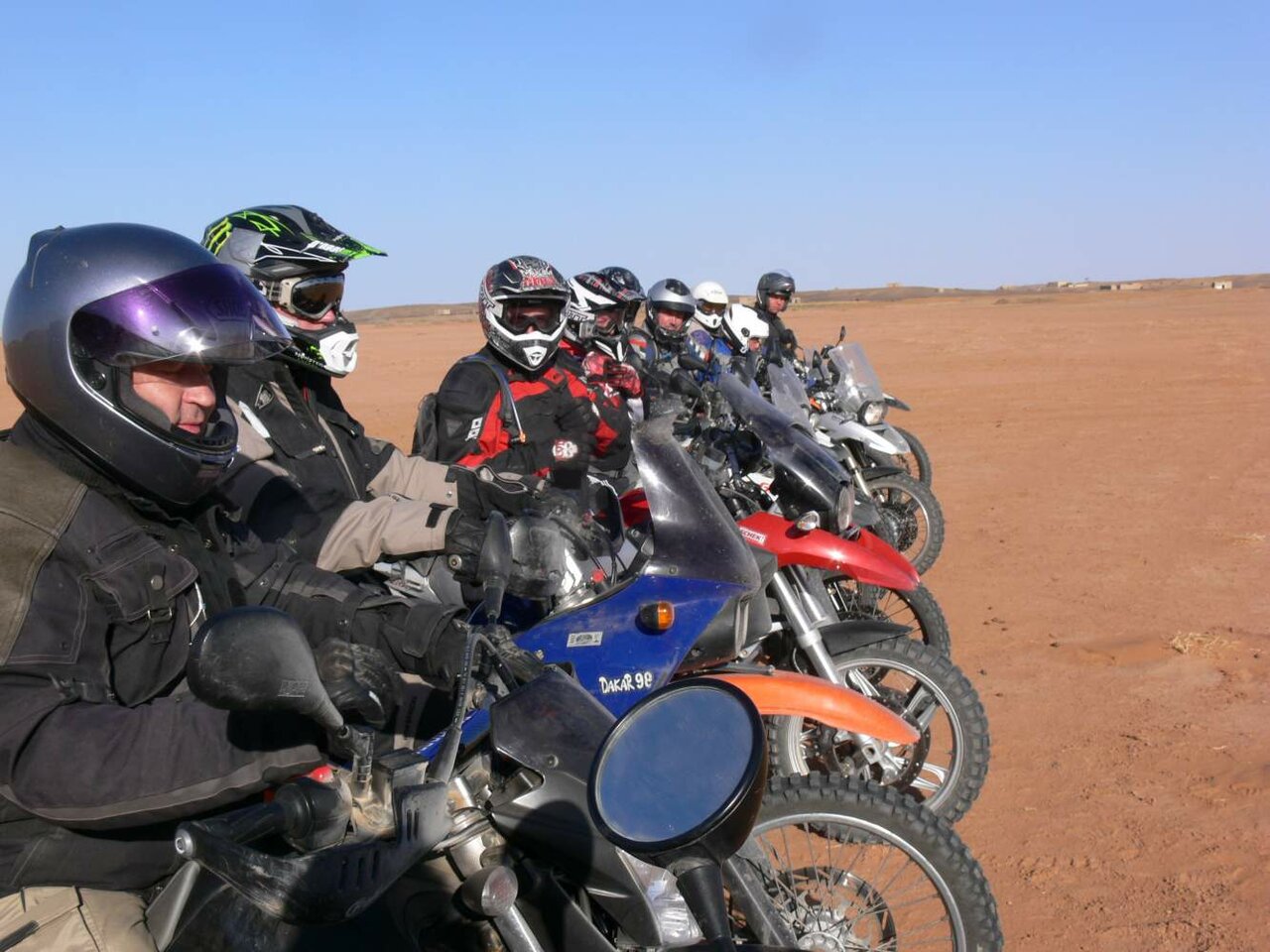 Fileira de pilotos e motos de trilha alinhados numa planície desértica plana, uma placa marcada Dakar 98