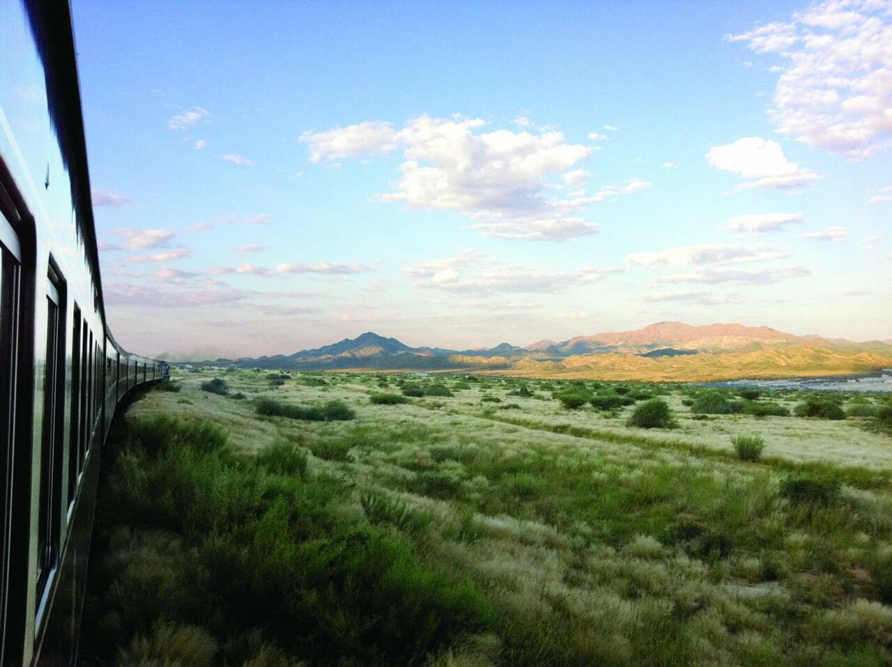 Vista de uma carruagem de comboio sobre vegetação rasteira verde com montanhas distantes e nuvens