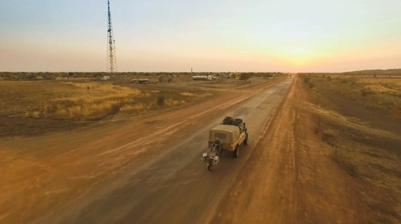 Luftaufnahme eines khakifarbenen 4x4 mit Dachgepäckträger und eines Motorrads auf einer Piste in der Dämmerung