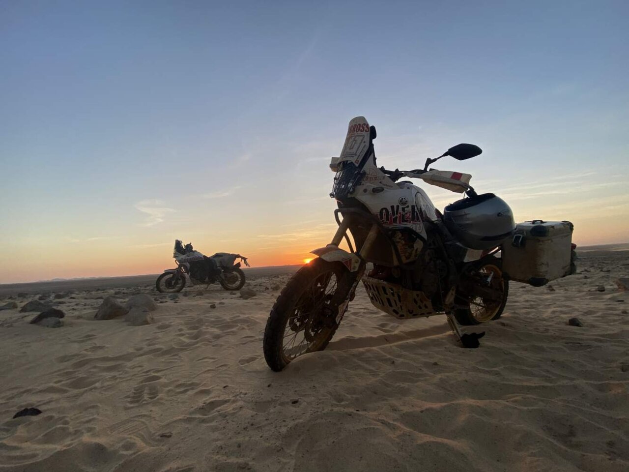 Silhuetas de três motos e dois pilotos com os braços erguidos numa crista do deserto ao pôr do sol