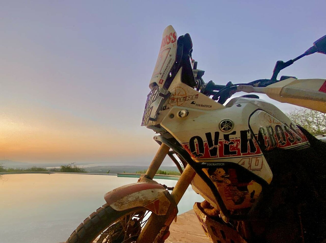 Uma moto de aventura coberta de pó, junto a uma piscina infinita com vista para uma paisagem enevoada ao anoitecer