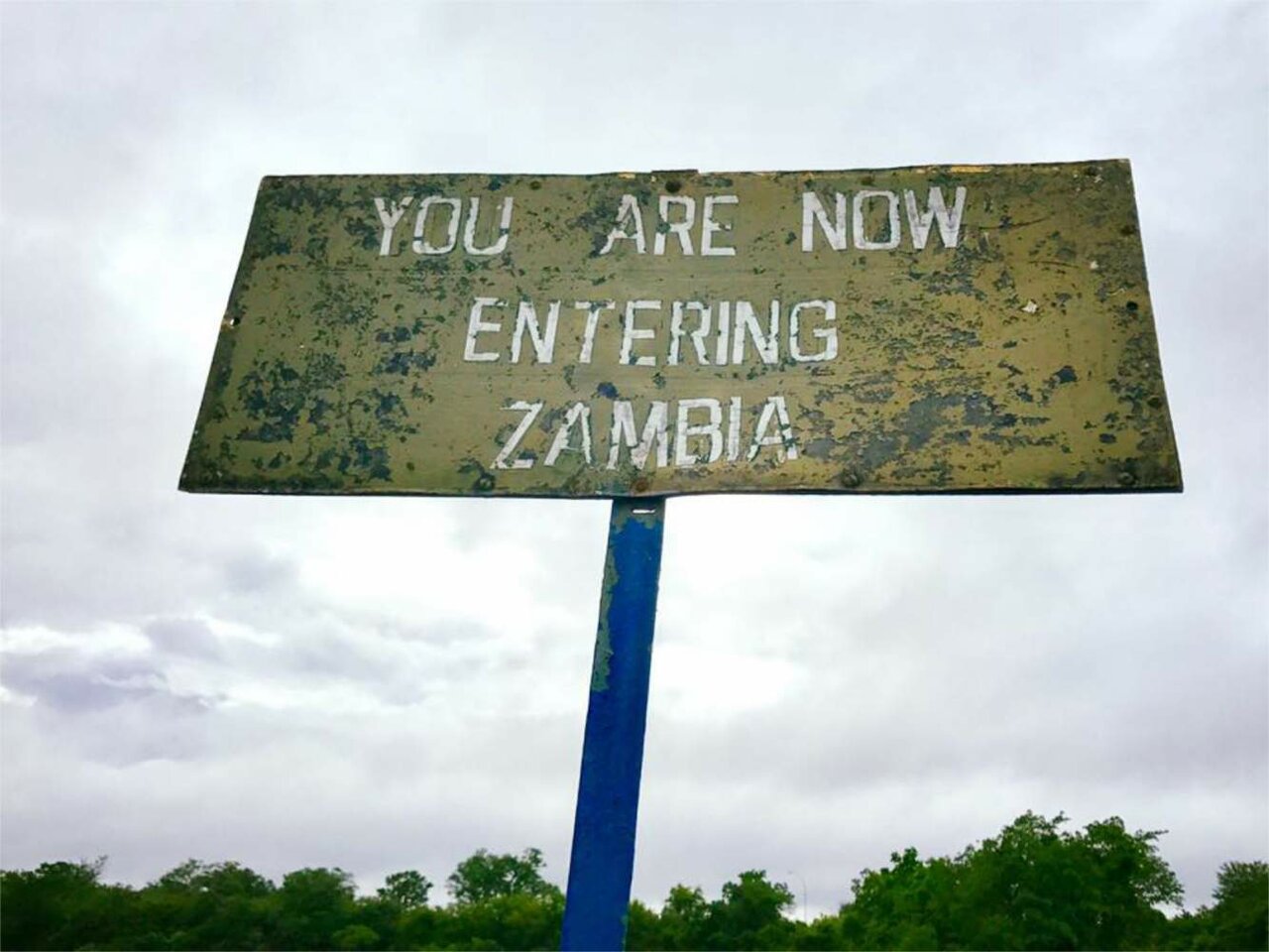 Un cartel desgastado junto a la carretera anuncia el paso fronterizo a Zambia bajo un cielo nublado