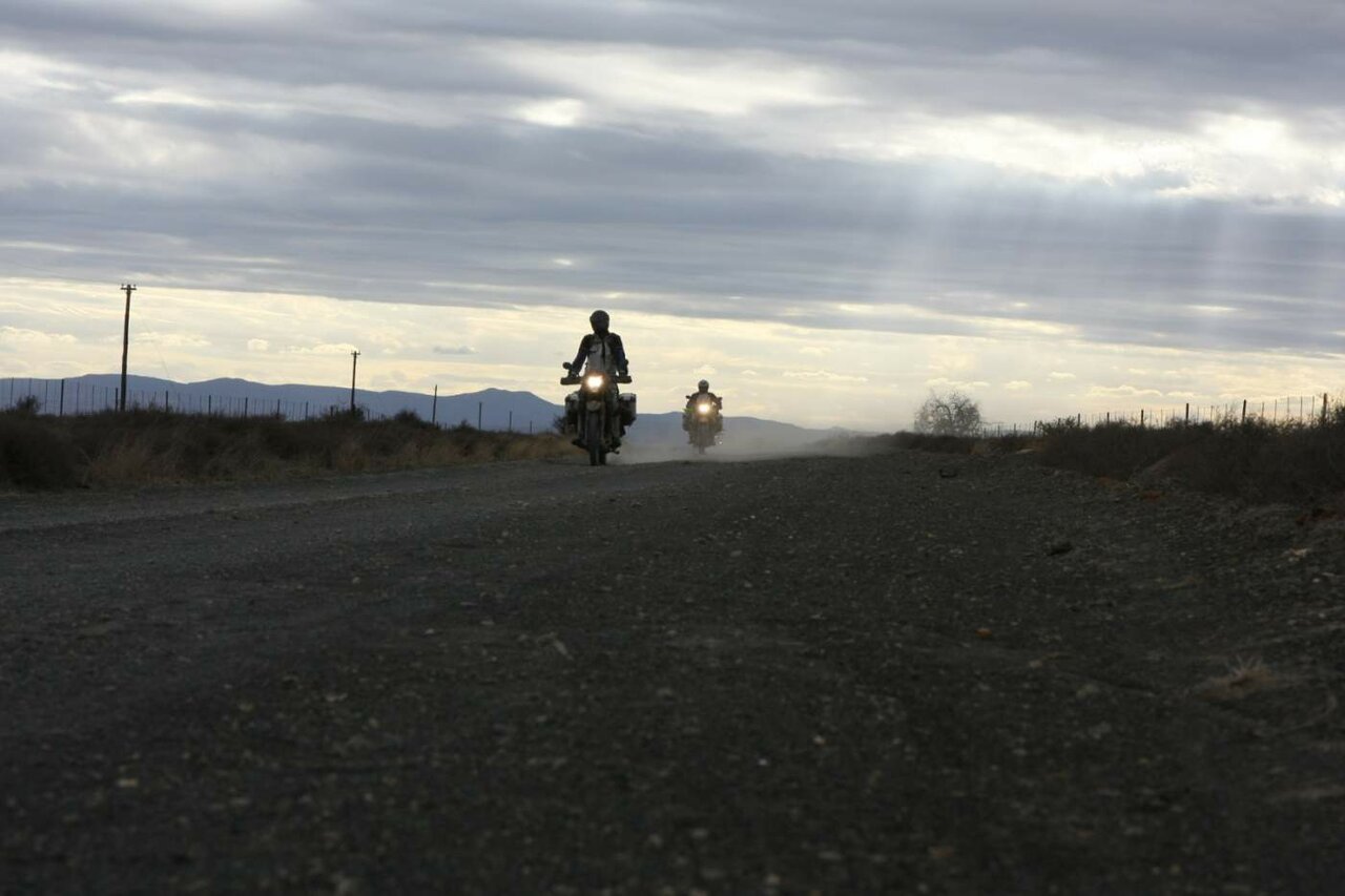 Zwei Motorradfahrer fahren in der Dämmerung eine Schotterstraße, Staub hinter ihnen