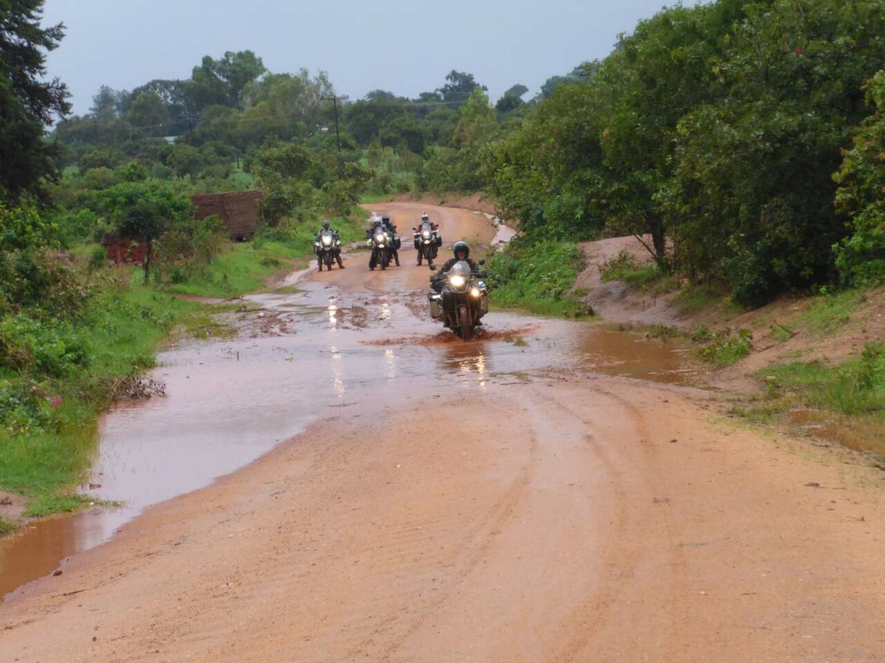 Motos atravesando un camino de barro inundado, salpicando agua entre árboles verdes
