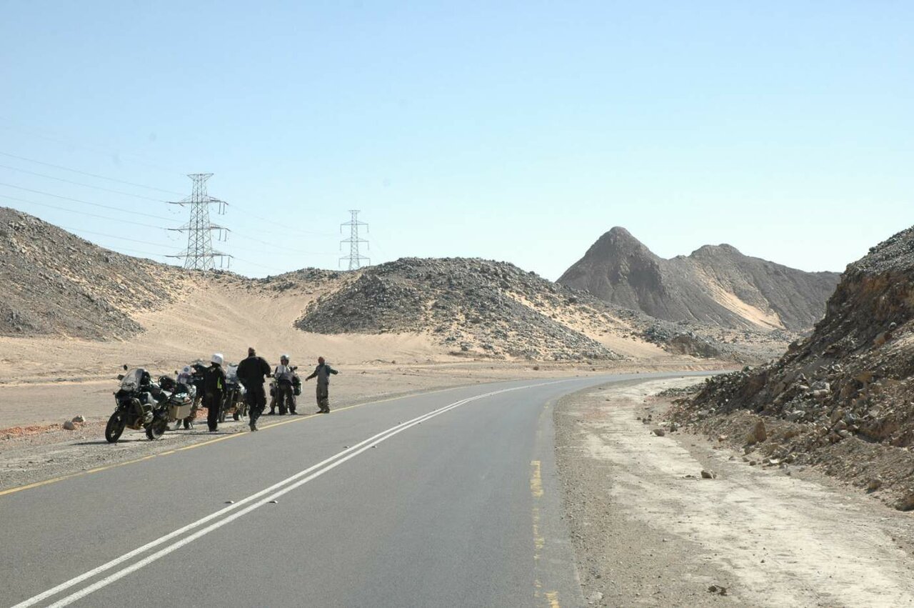Pilotos y motos parados junto a una carretera del desierto con colinas rocosas y tendido eléctrico