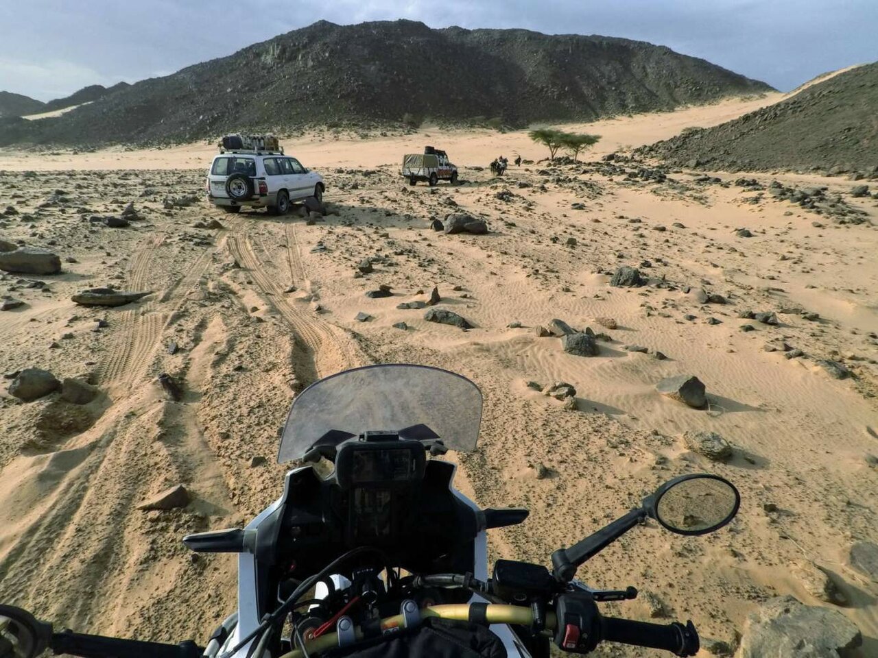 Vista desde el parabrisas de una moto sobre un desierto rocoso hacia dos vehículos de apoyo aparcados