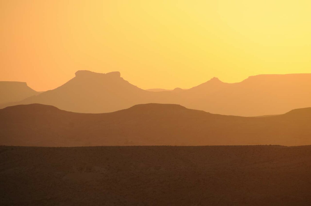 Gestaffelte Wüstenbergsilhouetten verschwimmen im Dunst unter einem goldenen Sonnenuntergangshimmel