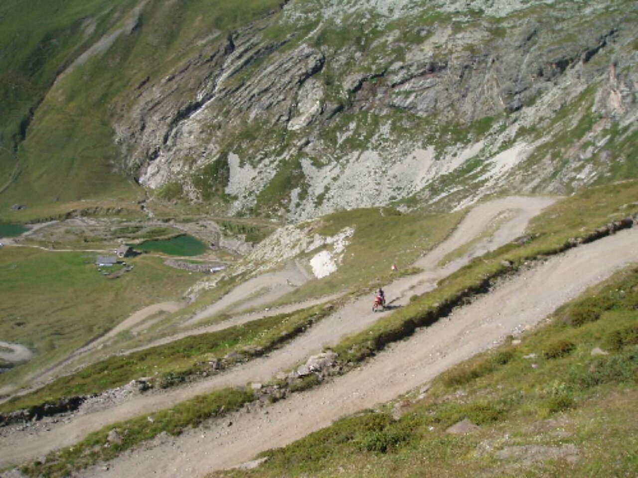Um motociclista distante numa trilha de terra acima de penhascos, com pequenos lagos no vale abaixo