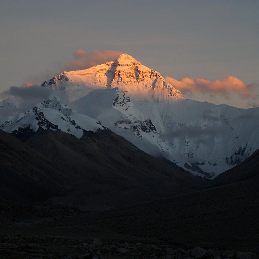 Ein schneebedeckter Himalaya-Gipfel leuchtet bei Dämmerung orange über dunklen Vorbergen