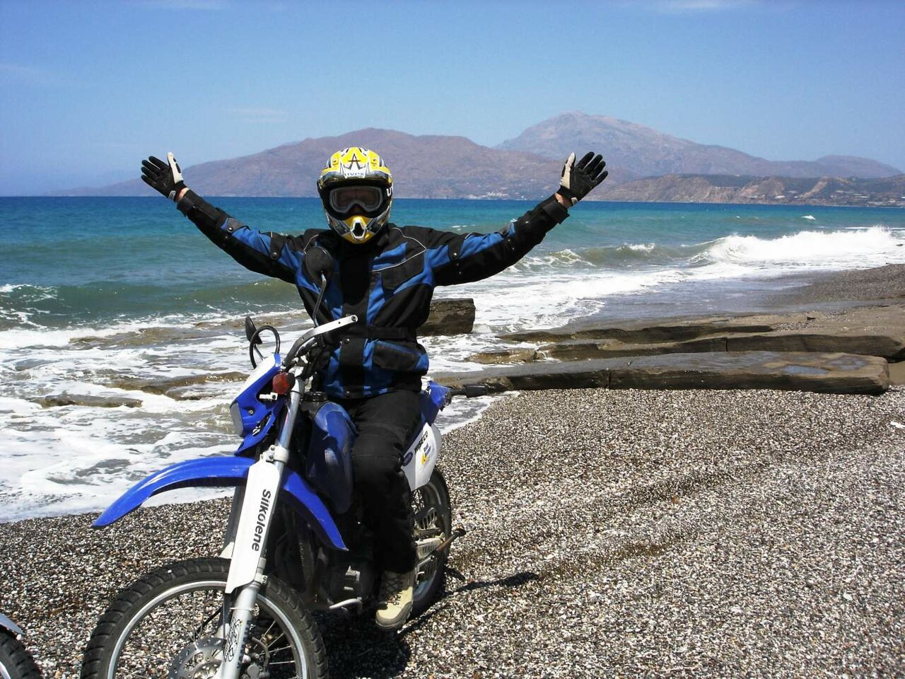 Ein Fahrer streckt auf seinem Motorrad beide Arme hoch, an einem Kiesstrand auf Kreta, Meer und Berge dahinter