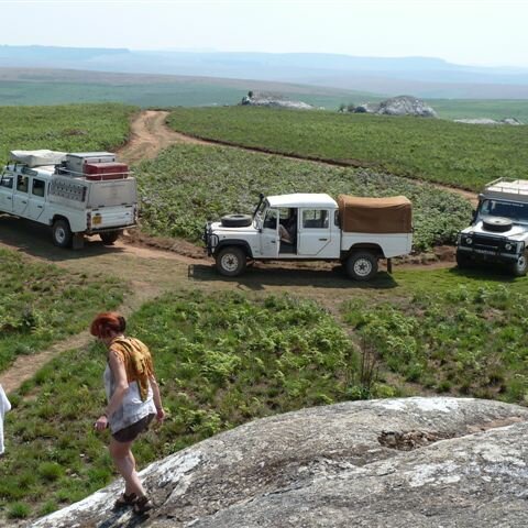 Drei 4x4 Safarifahrzeuge auf einer Piste durch grüne Hügel, im Vordergrund klettern zwei Wanderer auf einen Granitfelsen