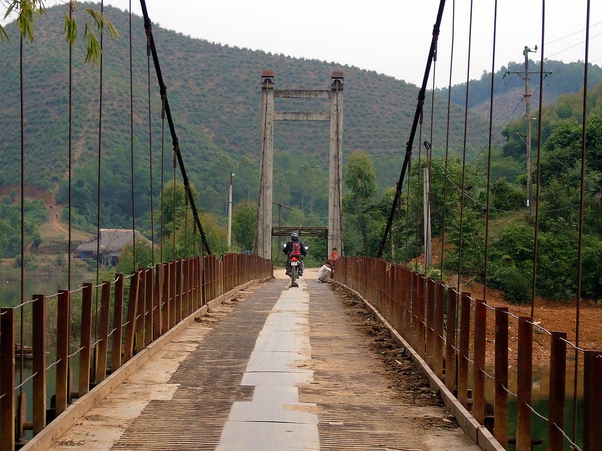 Motociclista atravessa uma ponte suspensa de aço, junto a uma cabana de colmo nas colinas