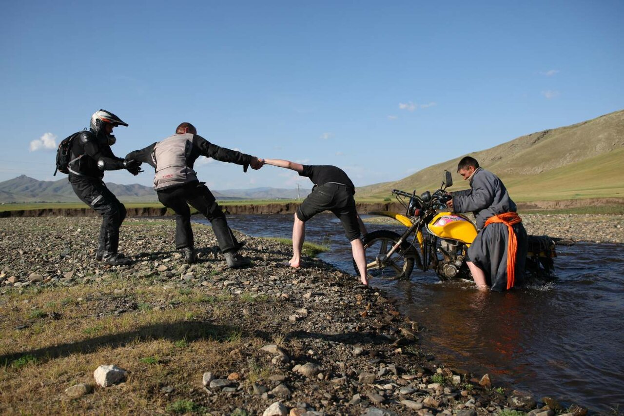 People forming a chain to help someone across a river beside a motorcycle standing in the water