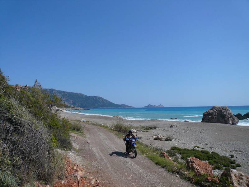 Enduro-Motorrad auf einer Schotterpiste an einer türkisfarbenen Bucht mit Felsvorsprüngen