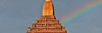 A tiered brick temple tower with a tall spire, greenery below and a rainbow in the sky