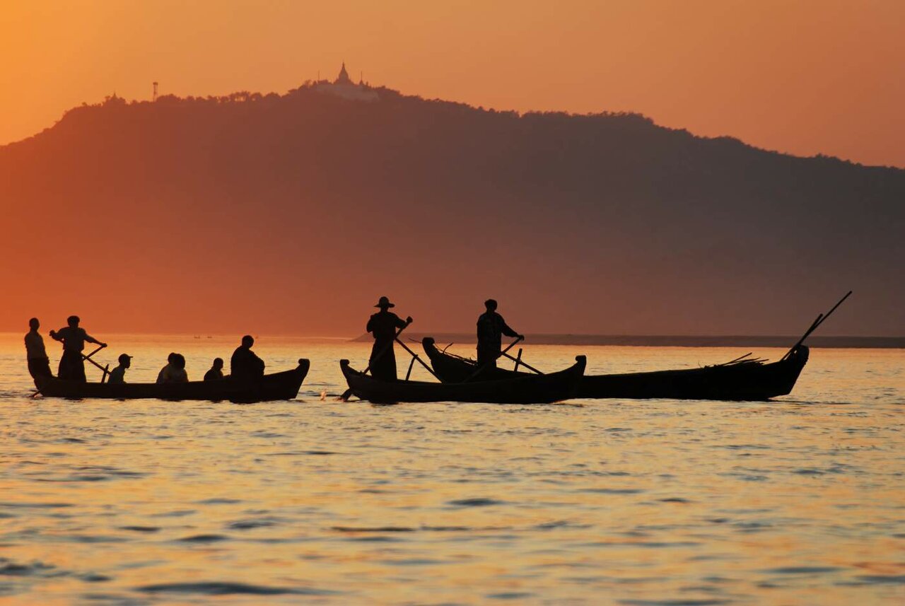 Silhouetted fishermen in boats on the Irrawaddy River at sunset, a hilltop pagoda in the distance