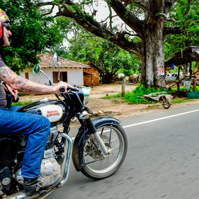 Ein Fahrer auf einer Royal Enfield an einem Dorfhaus und Straßenstand in Sri Lanka