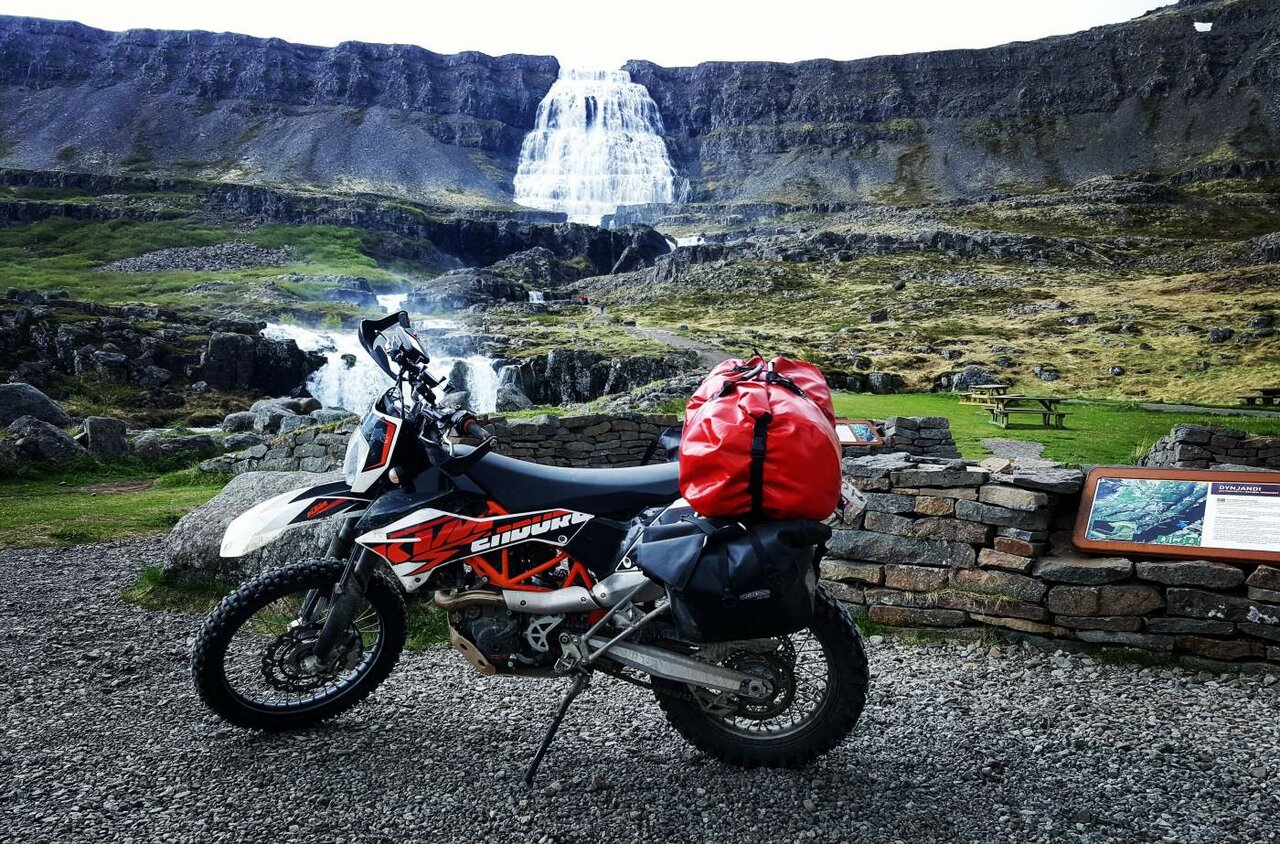 Ein KTM Enduro-Motorrad mit rotem Gepäck steht vor dem gestuften Wasserfall Dynjandi