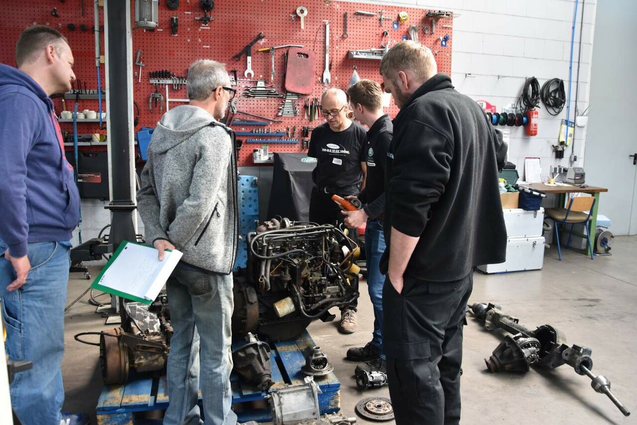 Gruppe untersucht ausgebaute Motor- und Achsteile auf einer Werkbank in der Werkstatt
