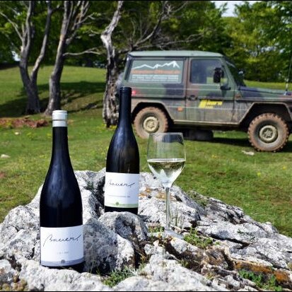 Zwei Weinflaschen und ein Glas auf einem Stein, dahinter ein schlammiges Offroad-Fahrzeug auf einer Waldlichtung