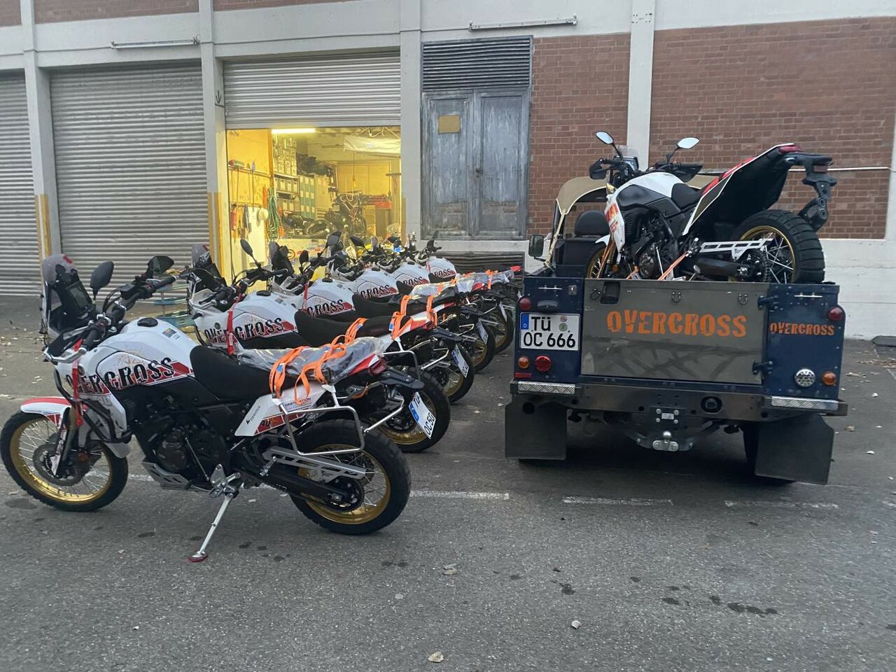 Adventure motorcycles lined up outside a garage, one loaded onto a pickup truck bed