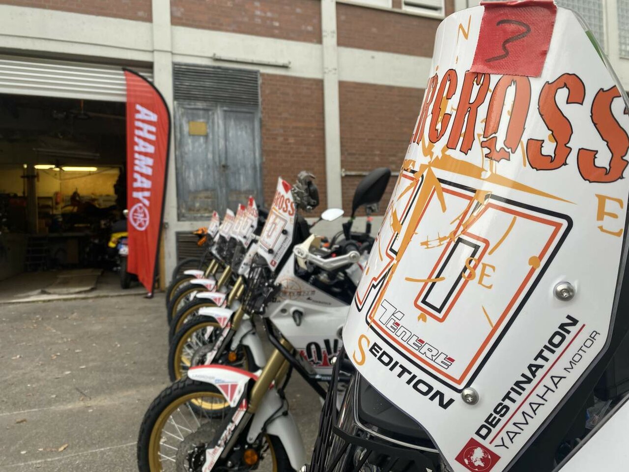 Eine Reihe Yamaha Tenere 700 Rally Mietmotorräder steht vor einer Werkstatt, mit Overcross-Aufklebern auf der Verkleidung