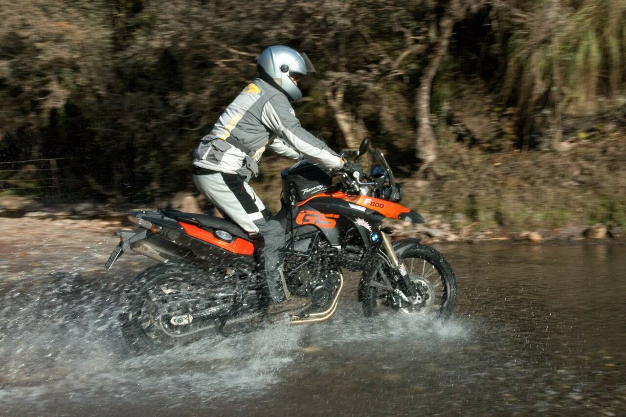Ein Motorradfahrer durchquert einen flachen Fluss neben einem bewaldeten Ufer