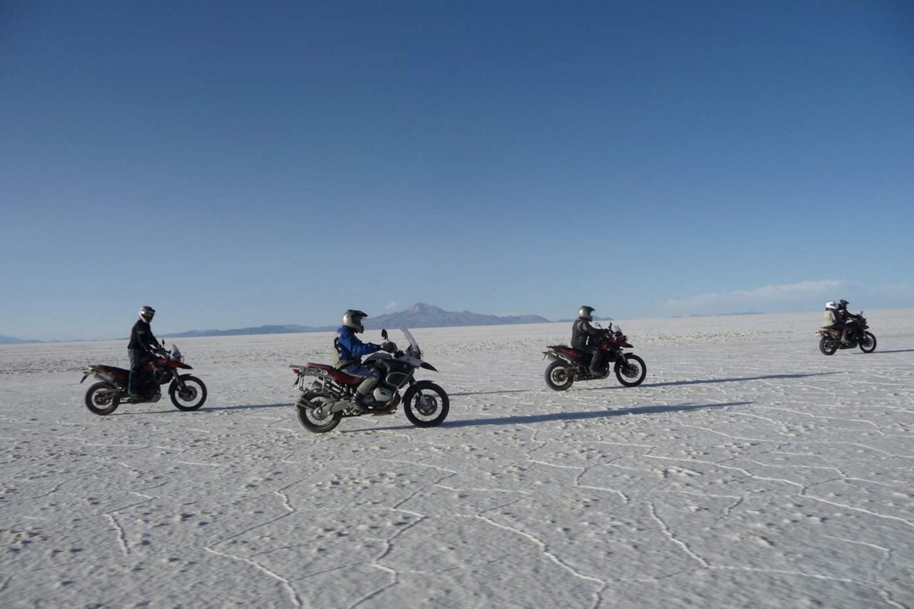 Dois pilotos descansam com uma moto numa encosta rochosa e alta, com um pouco de neve