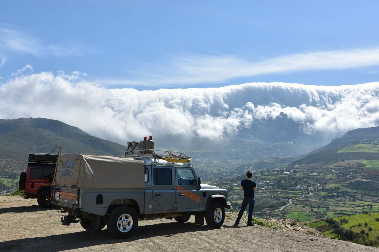 Ein 4x4 und ein Reisender auf einem Grat mit Blick auf Wolken über einem Bergtal