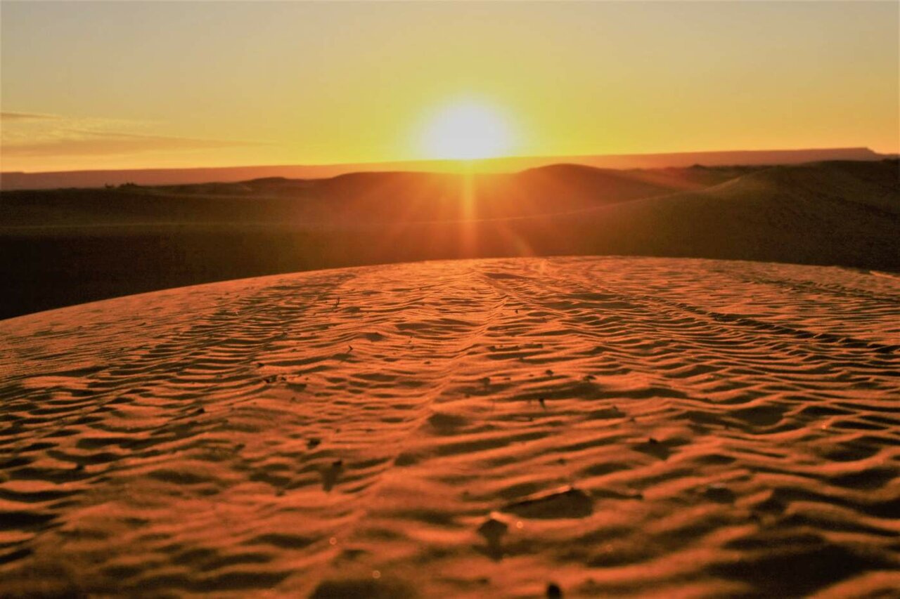 Die Sonne geht hinter Wüstendünen unter, Windrippel ziehen sich über den Sand im Vordergrund