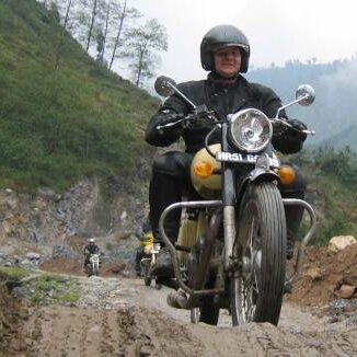Motorradfahrer auf Royal Enfields fahren eine steinige Piste hinab, dahinter dunstige Hügel