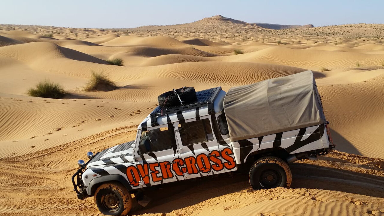 A zebra-striped 4x4 truck marked OVERCROSS parked on a Sahara sand dune