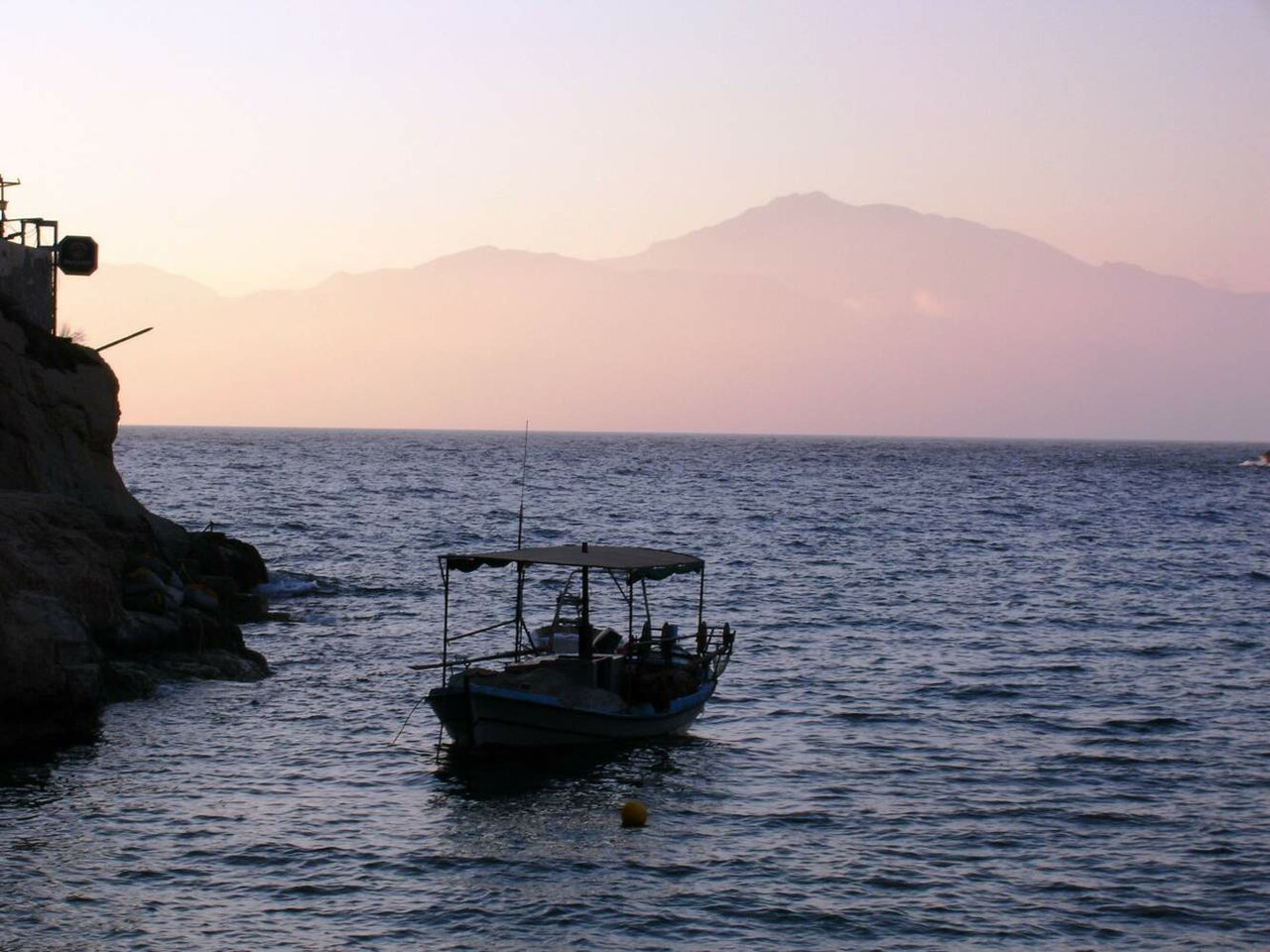 Small fishing boat moored near cliffs at dusk with hazy mountains across the water