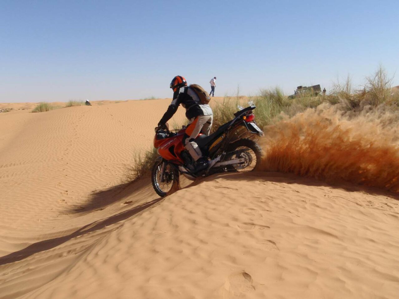 A dirt bike rider crosses a Sahara dune past tufts of desert grass with another rider ahead