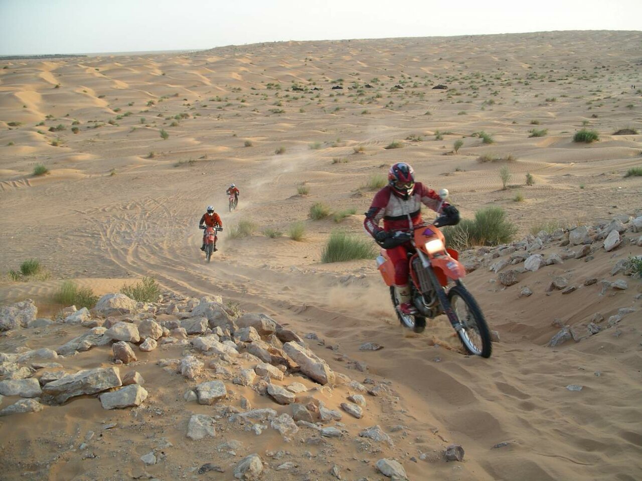 Drei Enduro-Motorradfahrer fahren auf einem staubigen Wüstenpfad an felsigem Boden vorbei