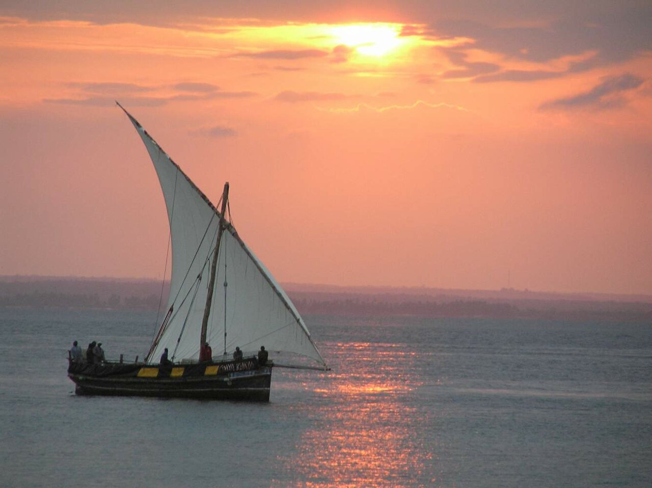 Ein traditionelles Dhau-Segelboot mit weißem Segel auf dem Meer bei Sonnenuntergang, Silhouetten an Bord