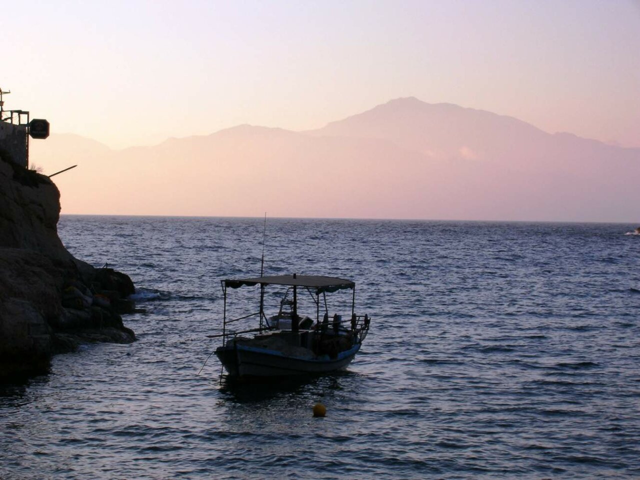 Silhouette eines vor Anker liegenden Bootes vor einer felsigen Küste unter rosafarbenem Abendhimmel