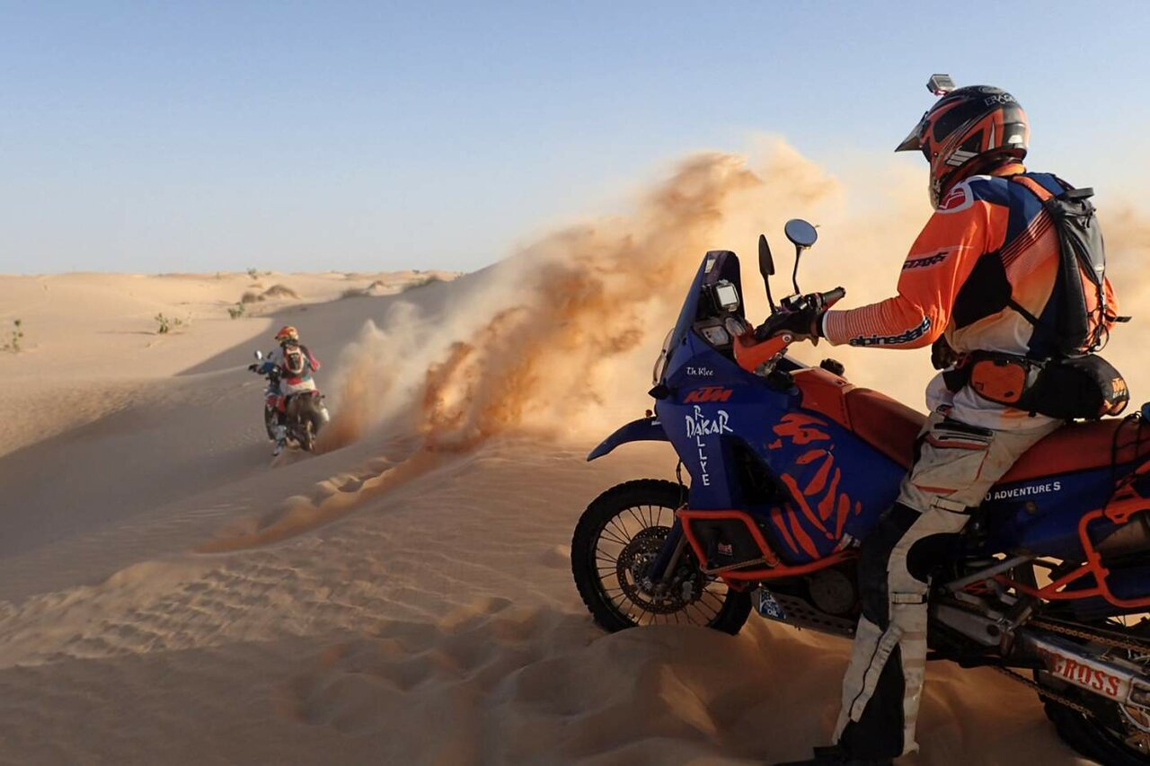 A rider on a KTM Dakar rally motorcycle watches another rider spray sand off a dune