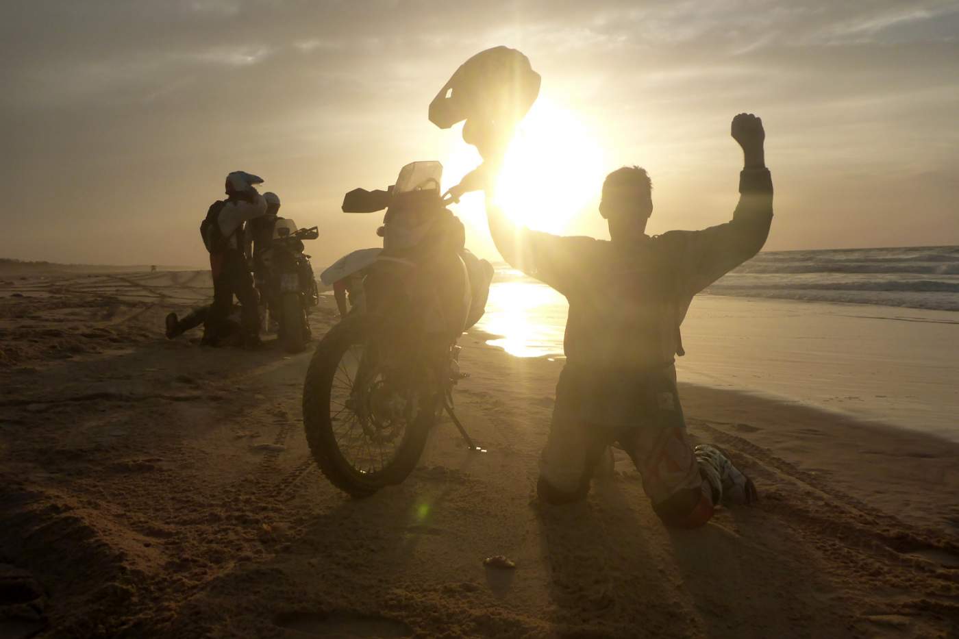 Motorradfahrer kniet am Strand von Dakar und jubelt über das Erreichen des Ziels dieser Motorradreise