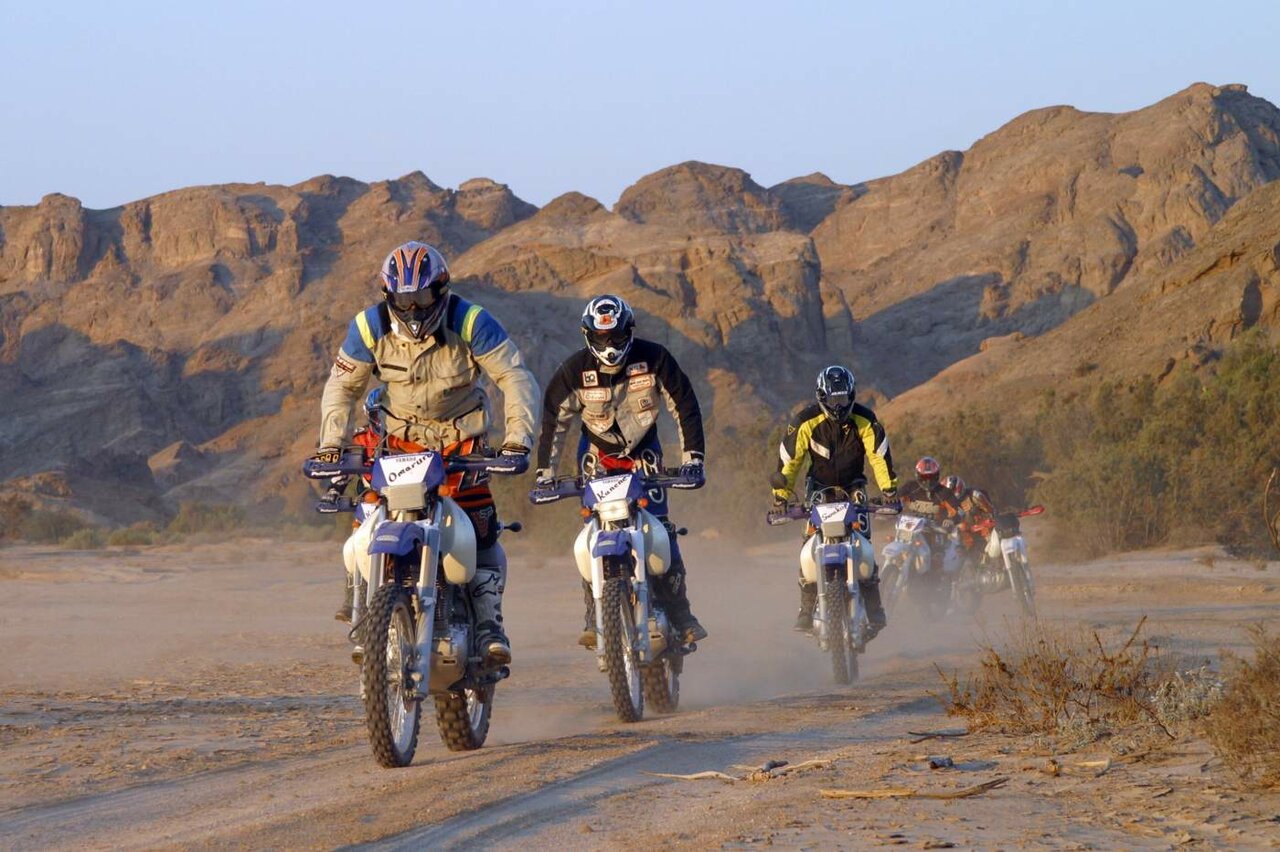 Mehrere Enduro-Fahrer durchqueren auf ihren Motorrädern eine staubige Schotterpiste unterhalb felsiger Wüstenberge