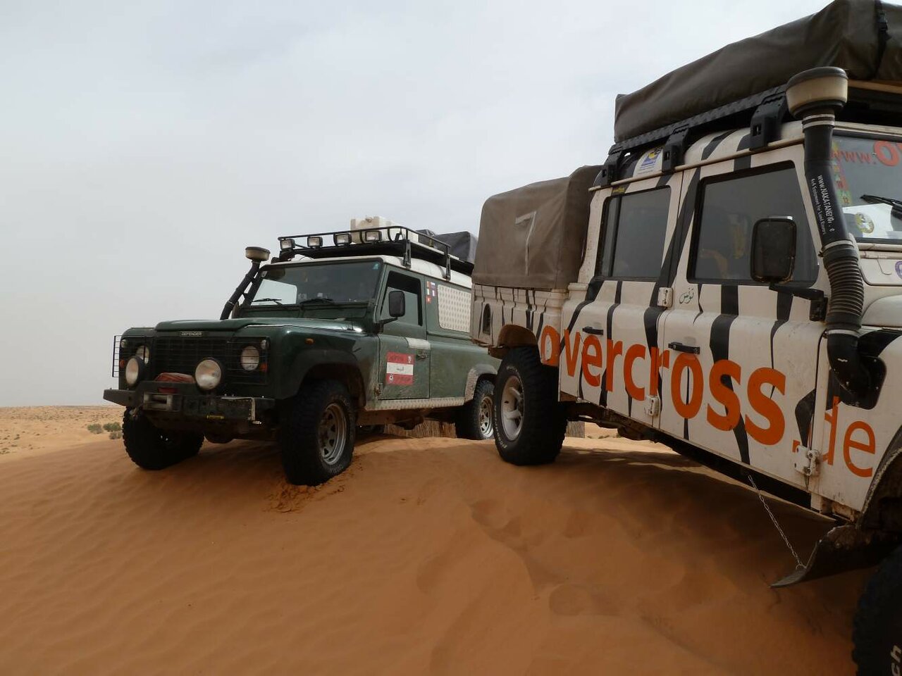 A green Land Rover Defender beside an overcross zebra-striped 4x4 on a sand dune
