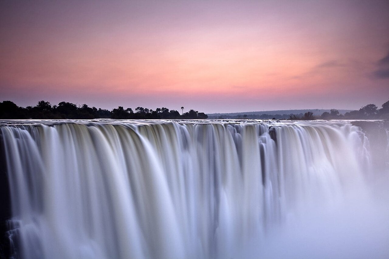 Victoria Falls bei Sonnenuntergang, ein breiter Wasservorhang stürzt in die Schlucht, Nebel steigt auf