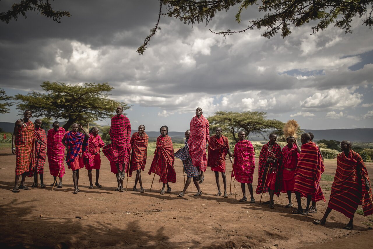 Eine Reihe Maasai-Männer in roten Shuka-Decken steht auf einer Piste unter stürmischem Himmel, einer springt in die Luft