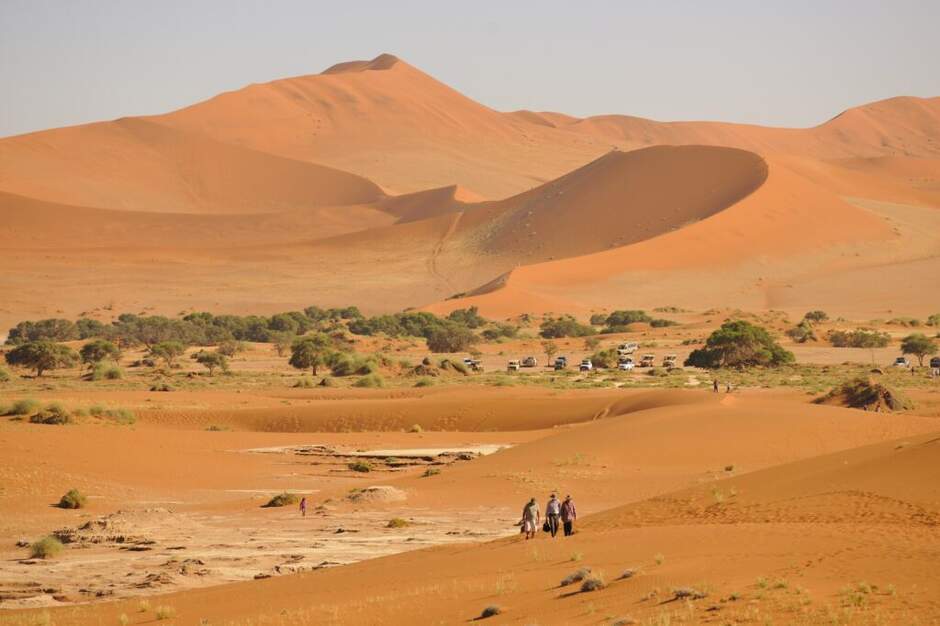 Hikers walking among red sand dunes with parked 4x4 vehicles in the valley below