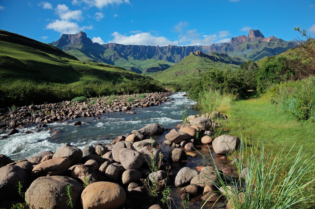 Um rio abaixo das colinas verdes e das falésias em camadas das montanhas Drakensberg