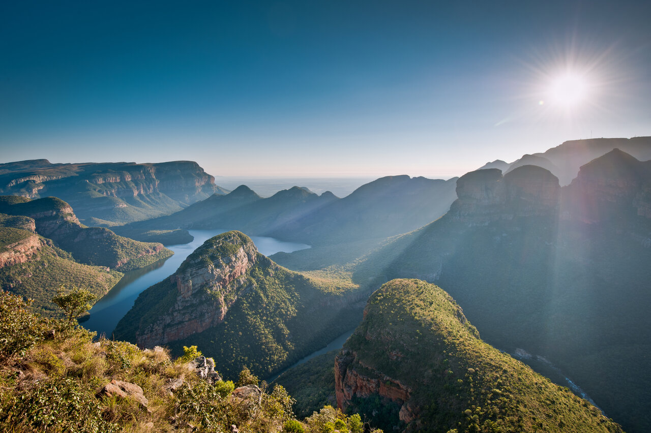 Um rio serpenteia por um canyon arborizado, com falésias de arenito em camadas e uma colina arredondada na neblina matinal